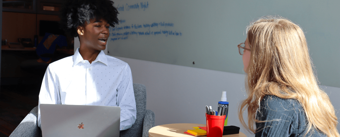 Two students sitting in chairs discussing a topic with a whiteboard behind them