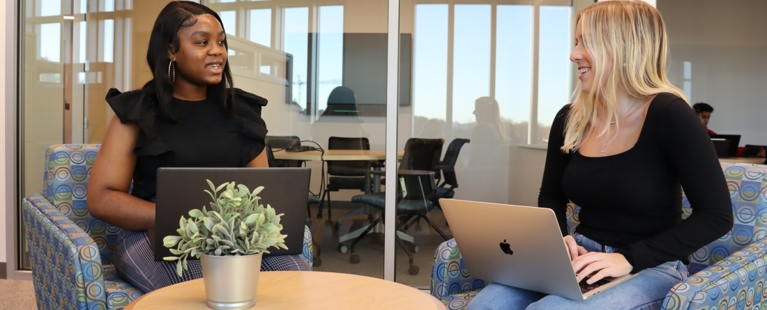 Two students sit in a chair with laptops conversing