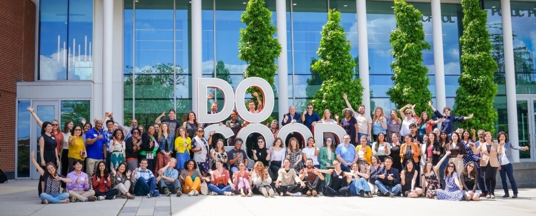 A large group of around 100 people pose in front of a glass building with four skinny green trees and a large sculpture that says Do Good. It is a nice blue and sunny day.