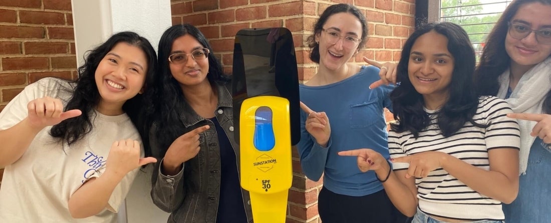 A group of five students pose beside and point at a yellow sunscreen dispenser. Red brick walls are in the background.