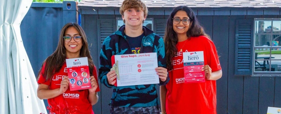 a woman with brown hair wearing a red shirt, a man with blnde hair wearing a blue shirt, and another woman wearing a red shirt all holding signs that say "Give hope" or "there goes my her." They are standing in front of a blue shed.