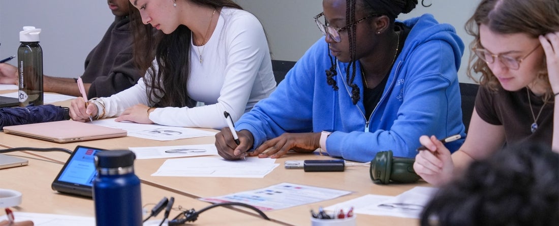 Four students sit at a beige colored table working and writing on a sheet of paper