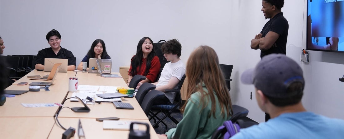 Students sitting at a long beige table listening to a man speak 